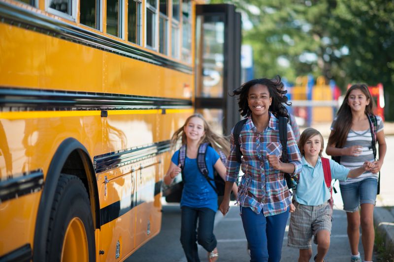 A multi-ethnic group of elementary age children are running from the bus to class and are smiling while looking at the camera.