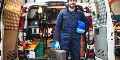 A smiling mechanic standing in front of an open van with tools, holding a toolbox and a helmet.