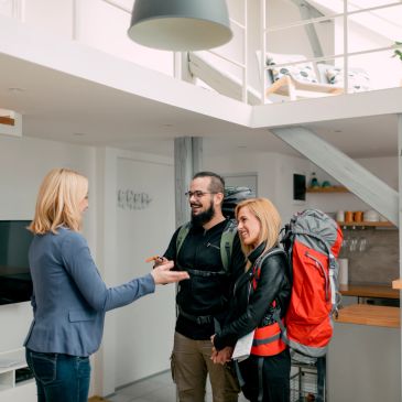 Couple with backpacks greeting a woman indoors.
