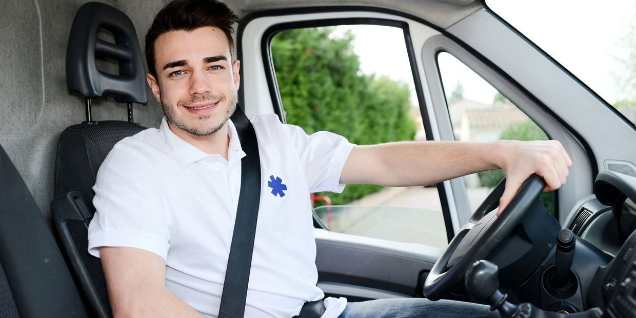 Smiling young man driving a vehicle, wearing a white polo shirt with a medical symbol.