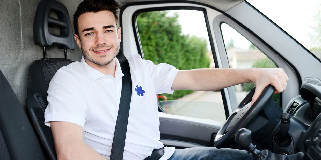 Smiling young man driving a vehicle, wearing a white polo shirt with a medical symbol.