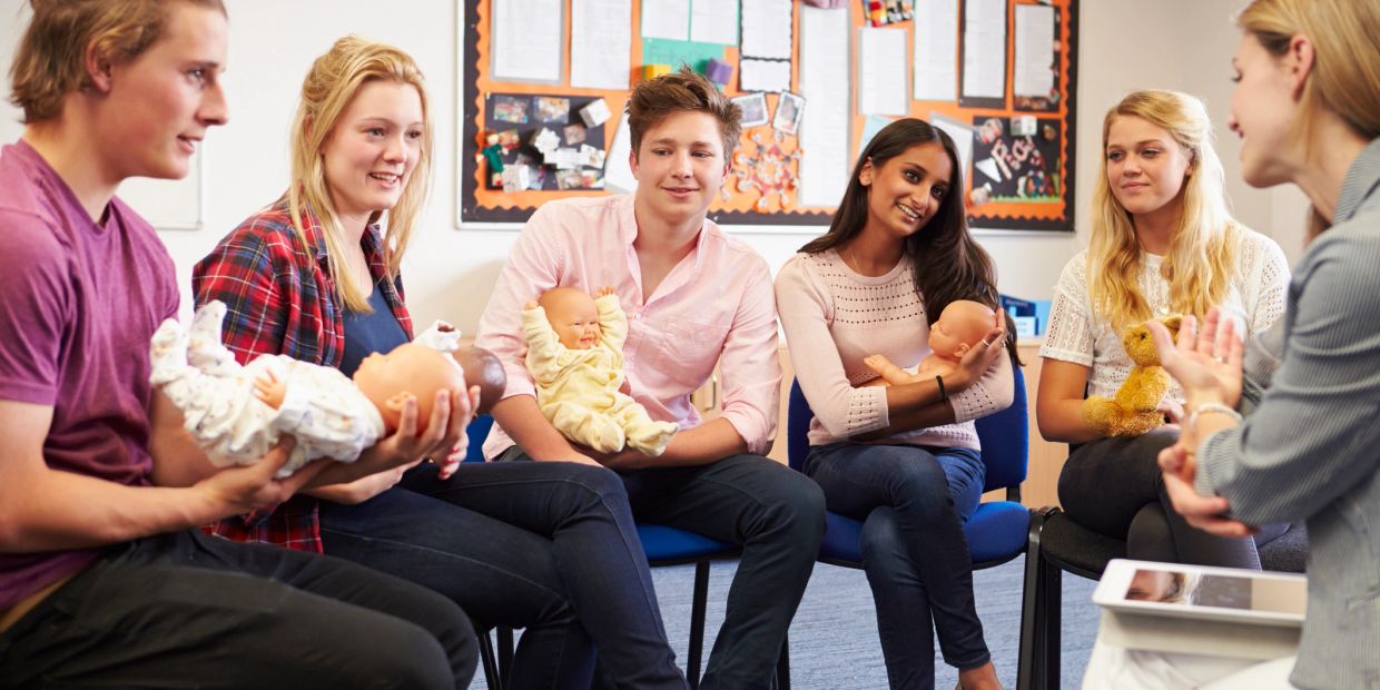 Group of young people holding baby dolls in a classroom.