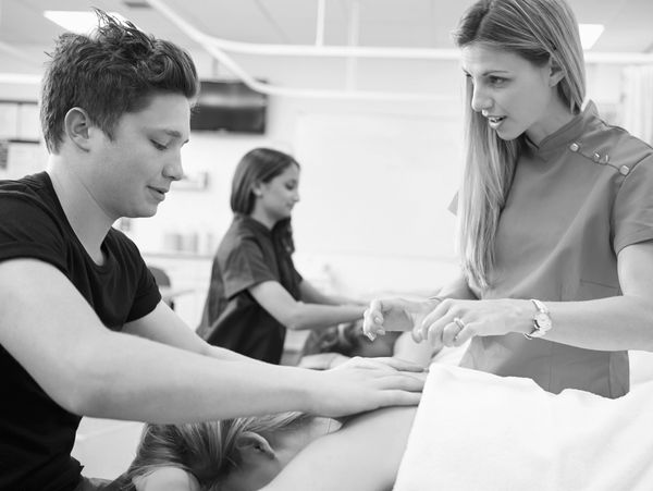 A massage instructor guides a young man practicing massage techniques on a client.