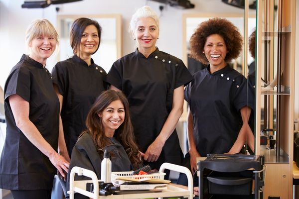 A group of smiling female hairstylists with a seated client in a salon.