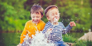 Two young boys joyfully splash water while sitting by a river.