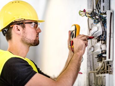 Electrician wearing safety gear testing electrical panel with multimeter.