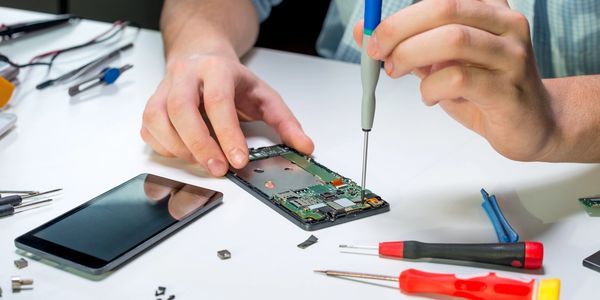 Person repairing a smartphone circuit board with precision tools on a white table.