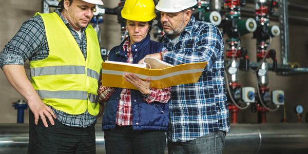 Three construction workers in helmets reviewing a manual in an industrial setting.
