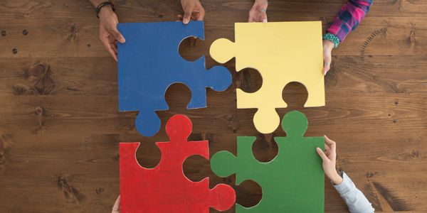 Four hands holding large colorful puzzle pieces over a wooden table.