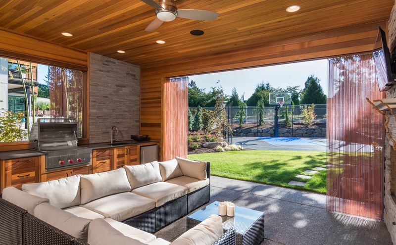 Covered patio behind new luxury home with fireplace, tv, and couch, with view of lush green grass and landscaping