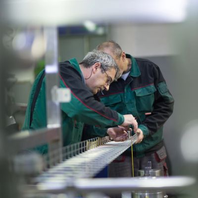 Two factory workers in green jackets inspecting a metal component.