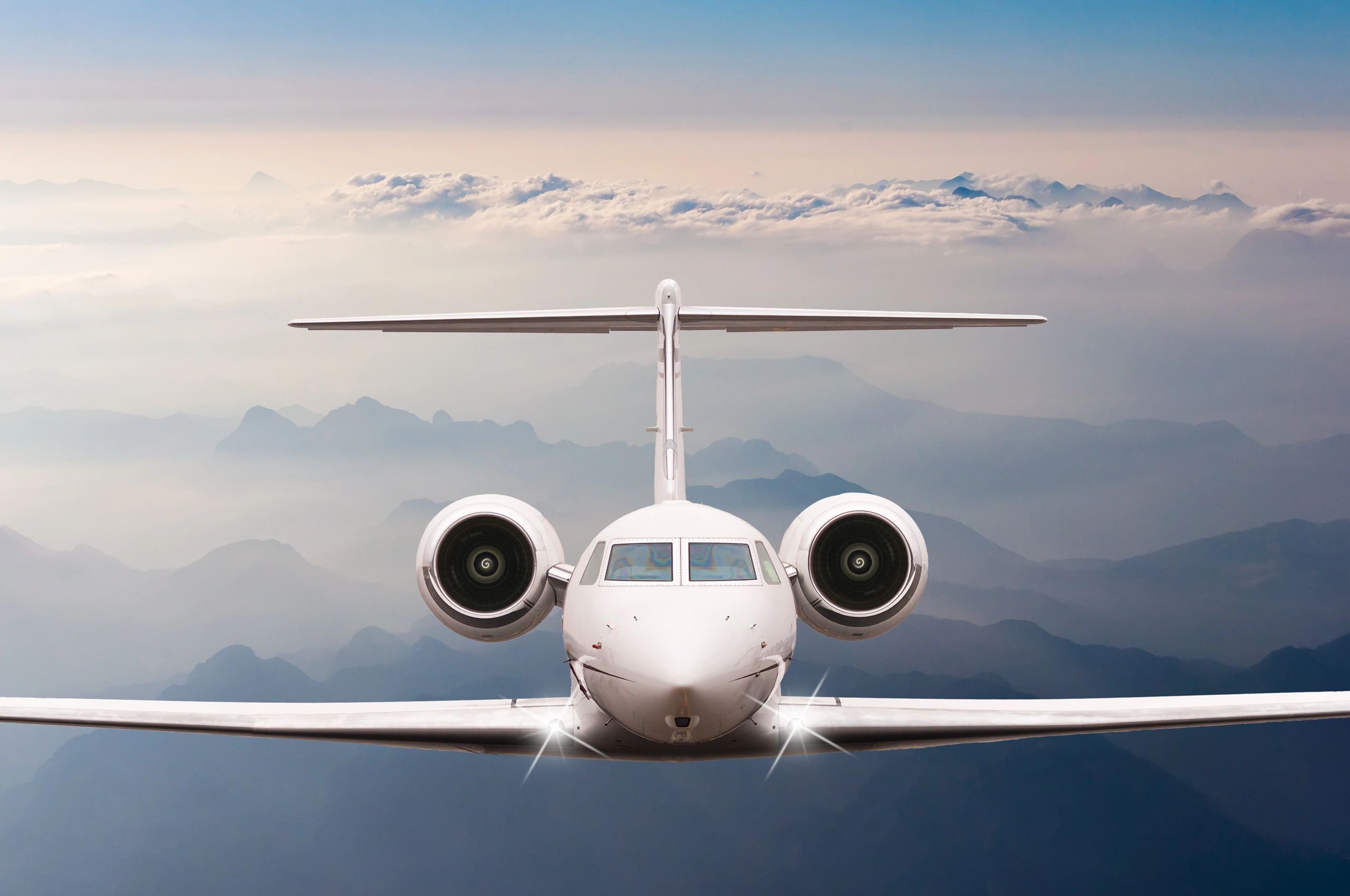Front view of a private jet flying above misty mountains and clouds.