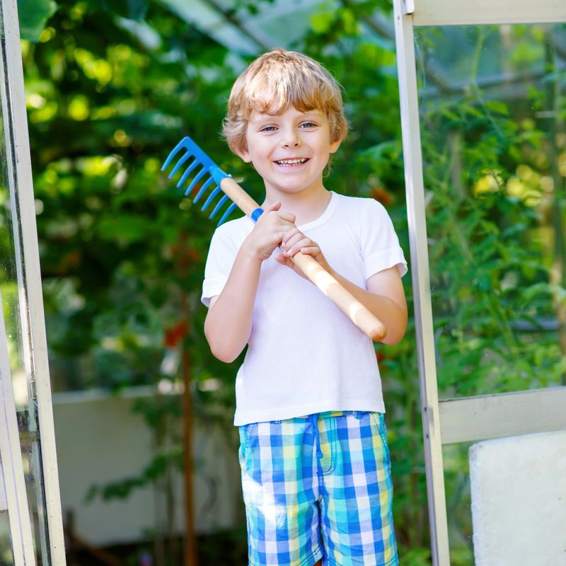 Smiling little kid boy watering plants and vegetables with can and working with garden hoe in greenhouse. Preschool child helping on sunny summer day. Family, garden, gardening, lifestyle