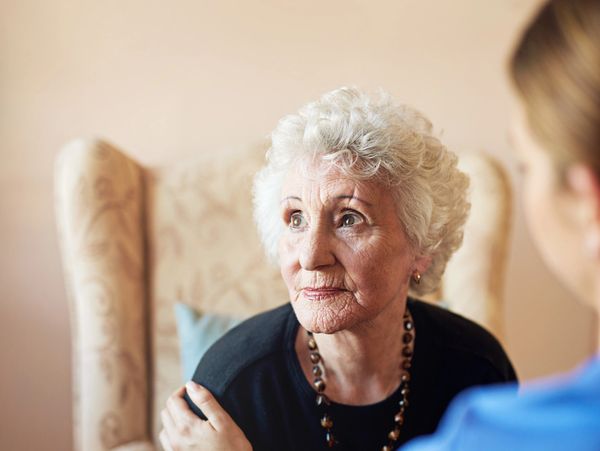 An elderly woman looks thoughtfully while being comforted by a caregiver.