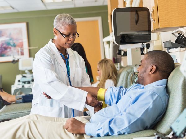 An African-American female doctor or nurse in a white coat and glasses is drawing blood