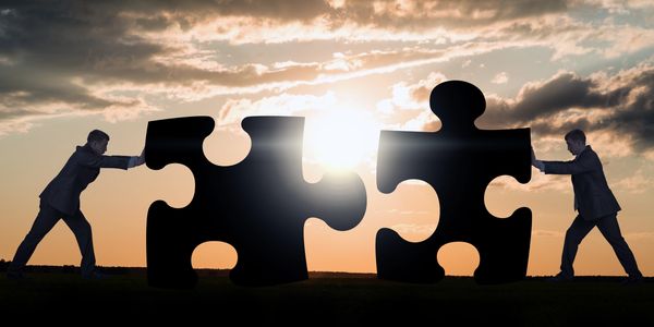 Two businessmen pushing giant puzzle pieces together at sunset.