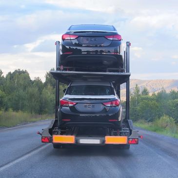 Two black cars stacked on a car carrier truck on a rural road.