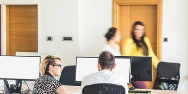 Two colleagues discuss work at computers while two others walk by in an office.