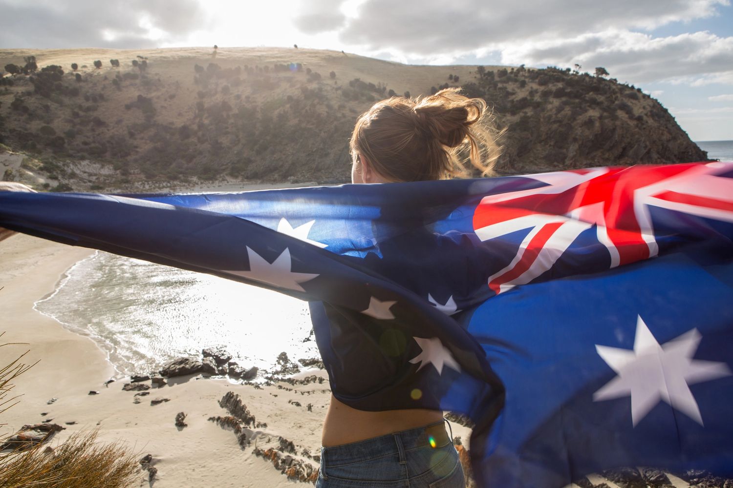 Woman with Australian flag overlooking a sunny beach and rocky cliffs.