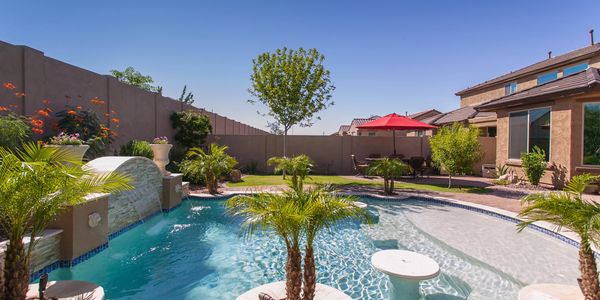 A backyard pool with palm trees, waterfall, and seating under a red umbrella.