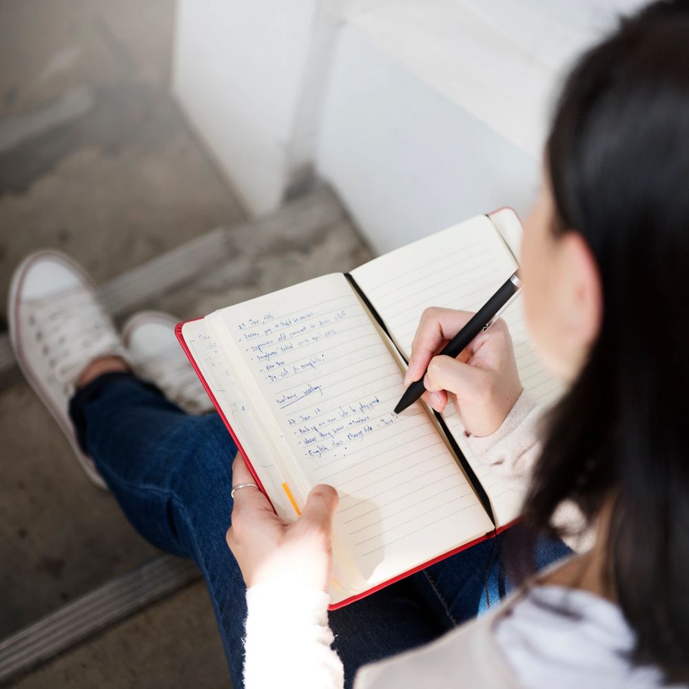 Person writing notes in a red notebook while sitting on stairs.