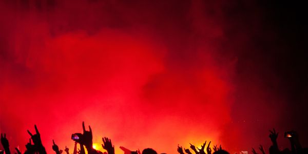 Silhouetted crowd raising hands against a fiery red background at a concert.