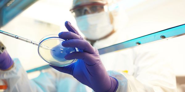 Scientist in protective gear using a pipette on a petri dish in a lab.