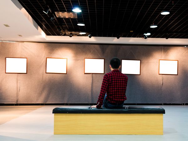 A person in a red plaid shirt sitting on a bench in an art gallery.