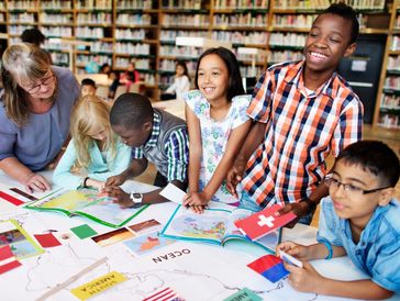 group of children & one adult looking at and researching atlases, world maps and flags.