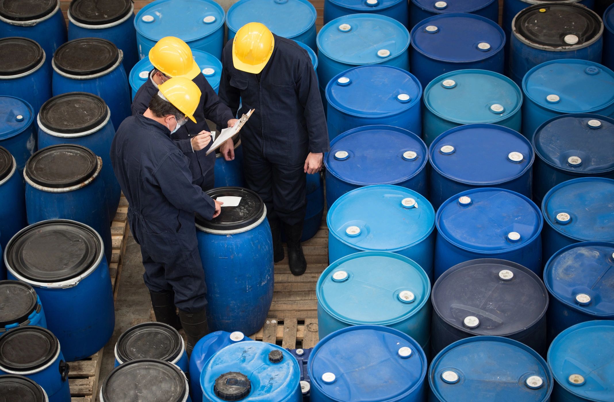 Workers inspecting blue chemical barrels wearing safety gear.