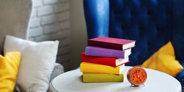 Stack of colorful books and an orange alarm clock on a white table.