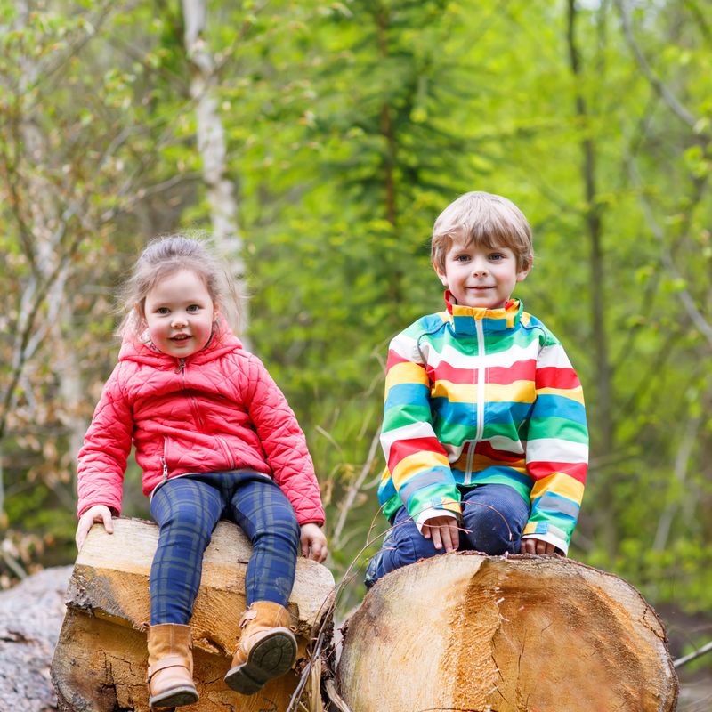 Two little cute smiling kids in bright jackets walking together in a forest on a rainy day. Friendship between siblings. Happy family concept