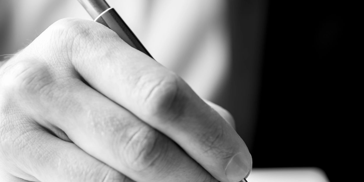 Close-up of a hand holding a pen, writing on paper in black and white.