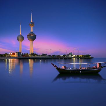 Traditional boat near Kuwait Towers at twilight with calm water reflections.
