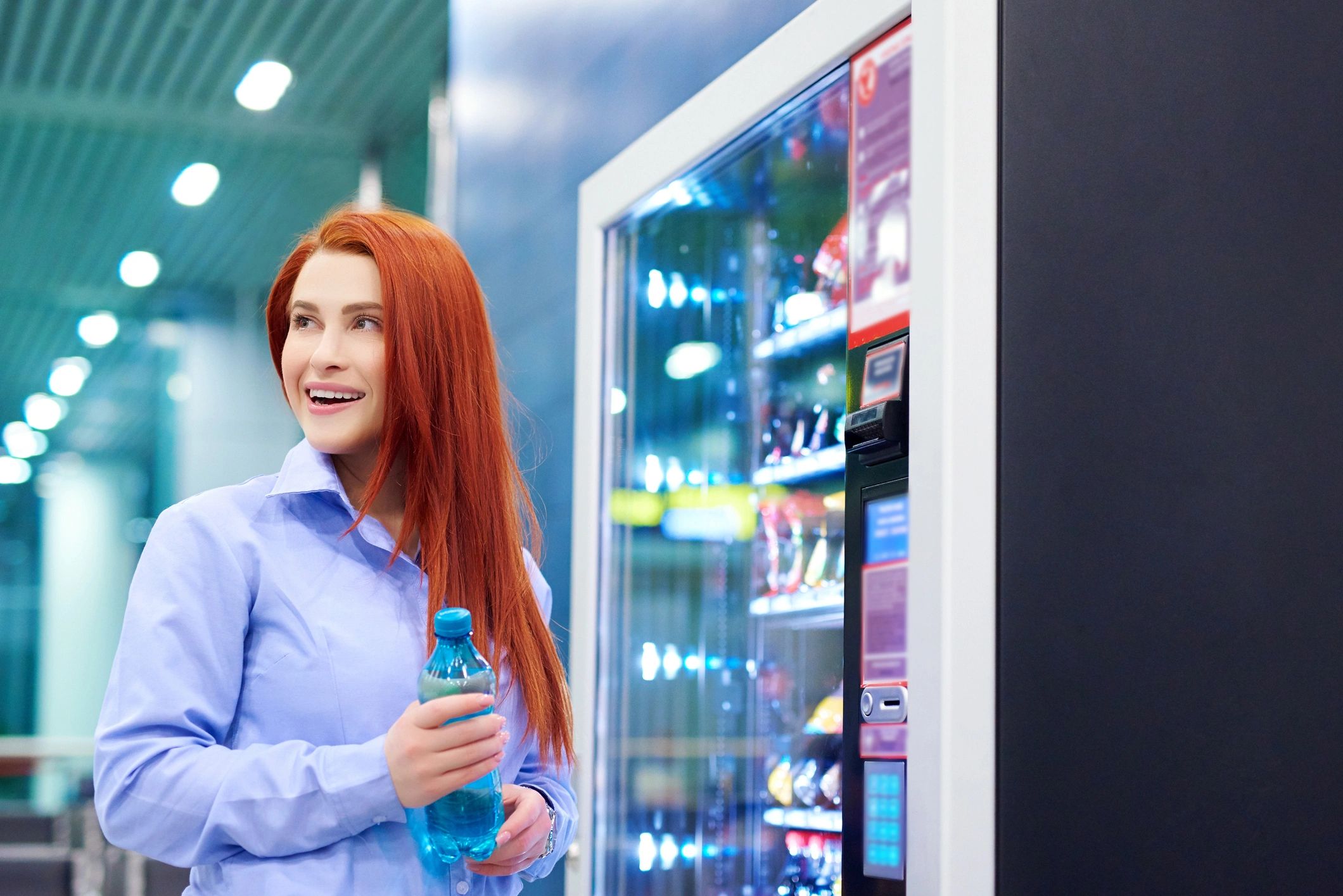 Smiling woman holding a bottle of water near a vending machine indoors.