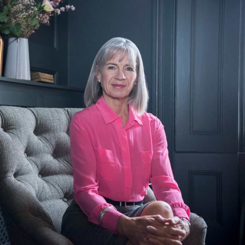 Senior woman wearing pink blouse sitting on grey armchair, looking at camera. Portrait of senior woman with grey bobbed hair.