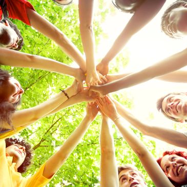 Group of friends joining hands in a circle outdoors under green trees.