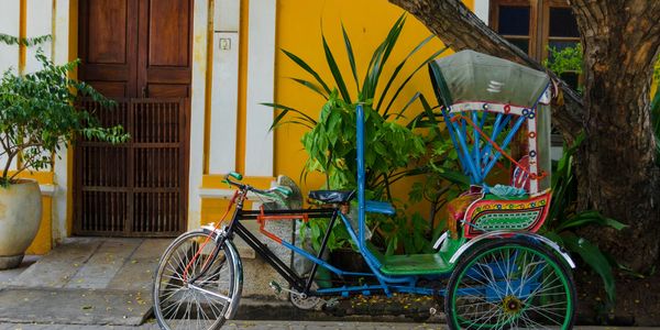 A colorful rickshaw parked beside a yellow wall with plants and wooden doors.