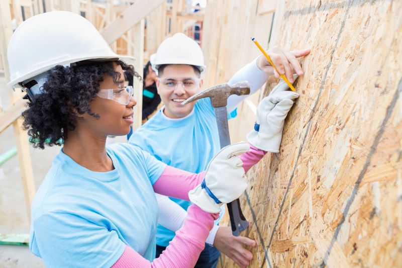 Mid adult African American female volunteer hammers a nail into plywood as mid adult Hispanic man marks the place for her. She confidently uses the hammer. The volunteers are working on building home for charity. They are wearing protective workwear.