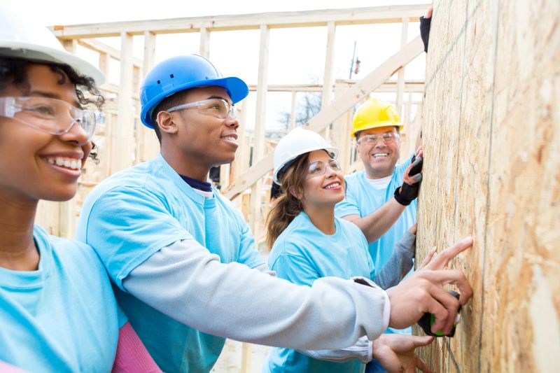 Diverse friends work together to raise wall in home they are building for charity. They smile confidently as they work on the wall together. They are wearing safety glasses, hard hats and light blue volunteer t-shirts.