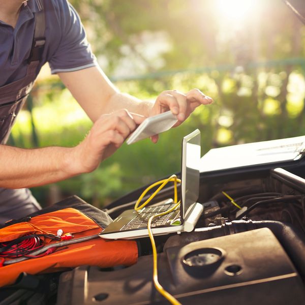 Mechanic using a smartphone and laptop to diagnose a car engine outdoors.