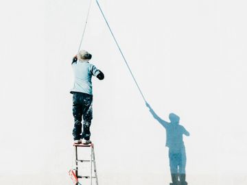 A man on a ladder painting a wall with his shadow mimicking the action.