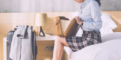 A woman in business attire sitting on a bed, looking into her purse with luggage beside her.