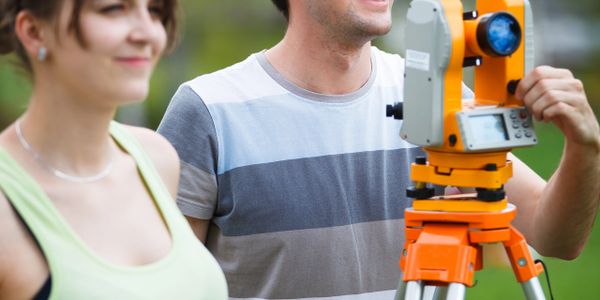 Two surveyors using a theodolite outdoors on a bright day.