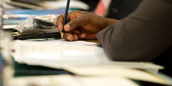 Person writing with a pencil on paper in a meeting or classroom.