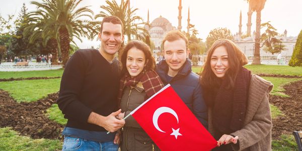 Four friends smiling and holding a Turkish flag in front of a mosque at sunset.