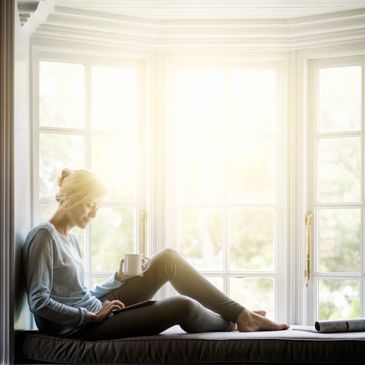 Woman relaxing with coffee and tablet by a bright window.