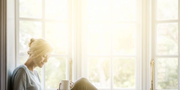 Woman relaxing with coffee and tablet by a bright window.