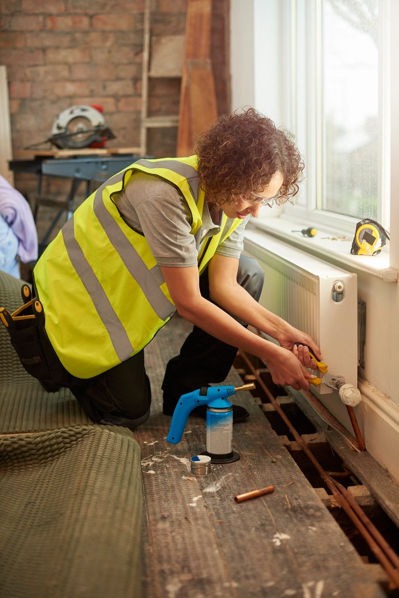 a mid adult female plumber is installing a new radiator into a property undergoing refurbishment. The floorboards are raised with new paperwork shown , and she is tightening the valve on the new radiator. She is wearing work trousers , and tool belt . In the background more work tools can be seen as is an exposed brick wall .