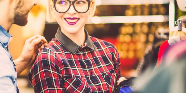 A young woman with glasses smiling while shopping for clothes with a companion.